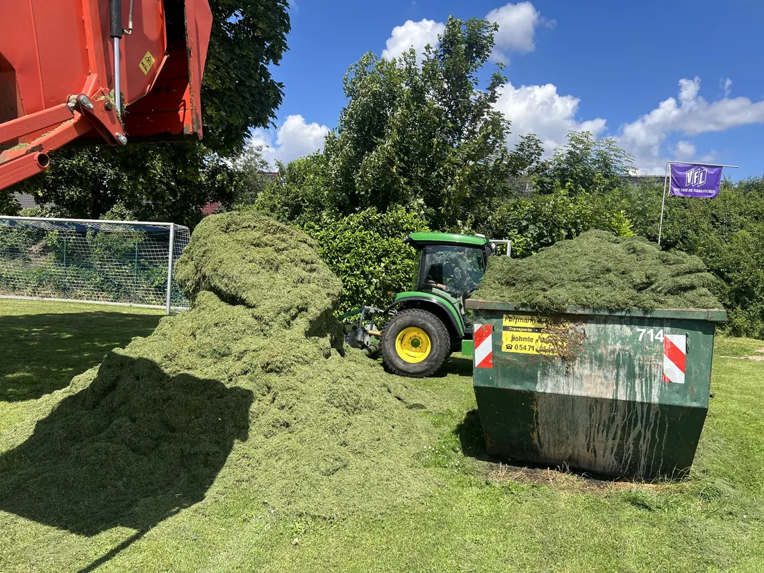 Gesammeltes Mähgut beim Entladen neben einem Container und einem John-Deere-Traktor.