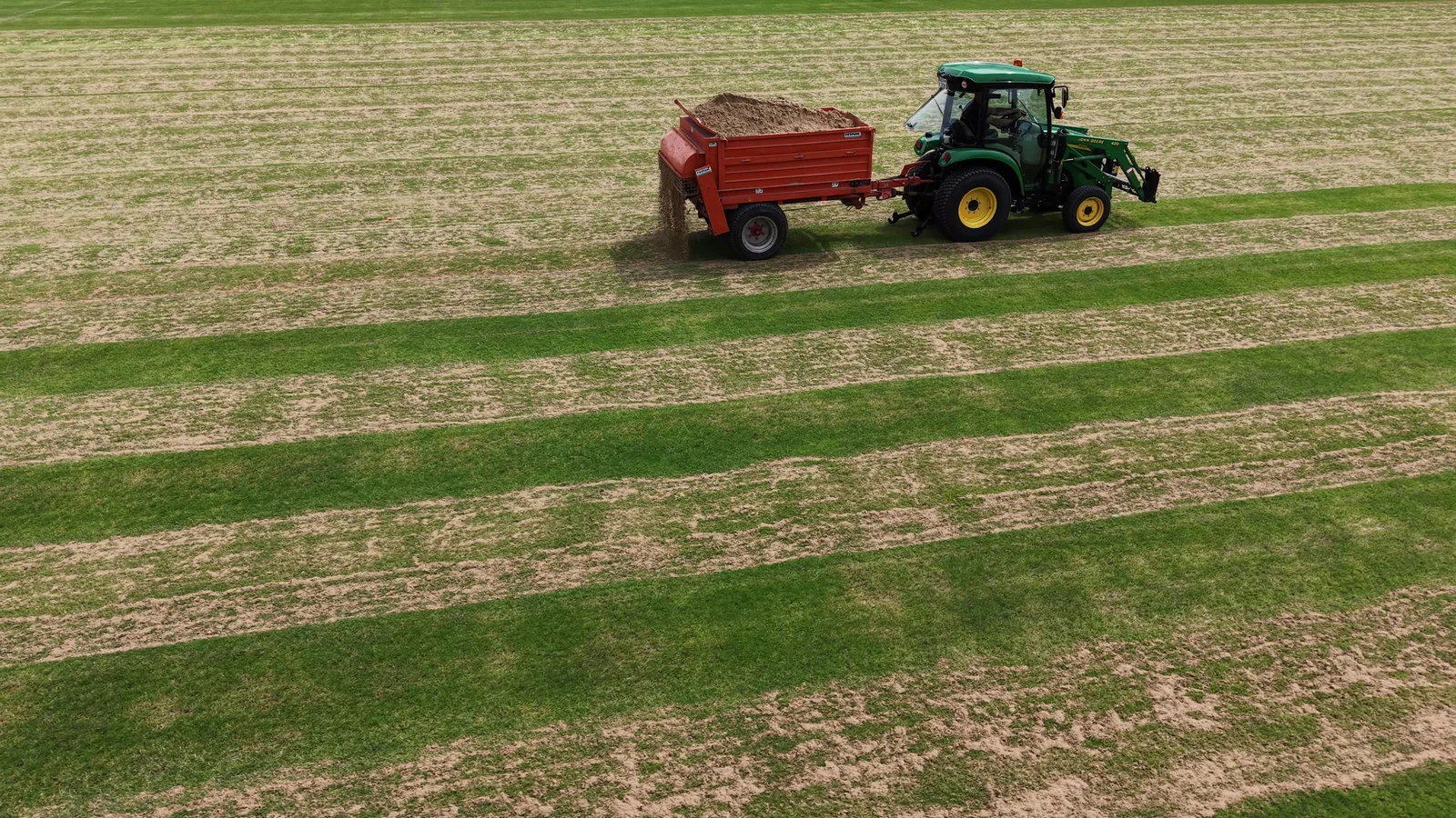 John Deere mit gezogenem Sandstreuer beim gleichmäßigen Besanden einer Naturrasenfläche.