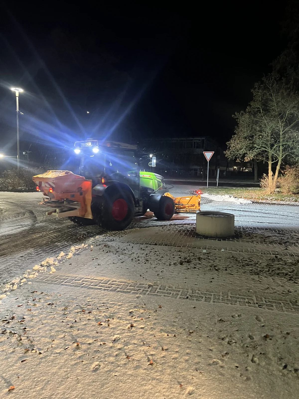 Traktor mit Schneeschild und Streuer beim nächtlichen Winterdienst auf einem Industriegelände.